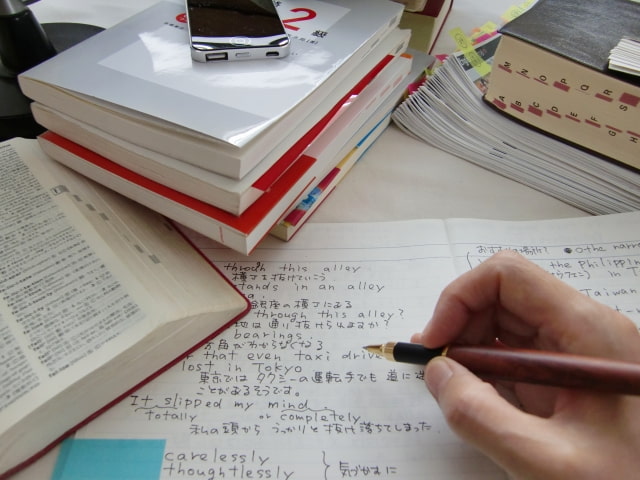 机上に英語の辞書や書籍を置き、右手で鉛筆を持ち、ノートに英文を書いている